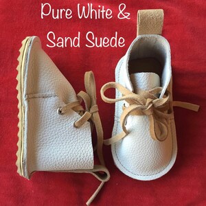 May include: A pair of white leather baby shoes with tan suede laces. The shoes have a textured leather upper and a white rubber sole. The text "Pure White & Sand Suede" is written above the shoes.