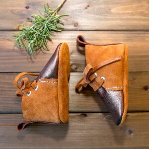 May include: Two brown leather baby shoes with laces. One shoe is brown with a brown leather toe and the other shoe is brown with a dark brown leather toe.