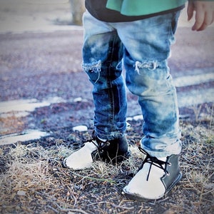 May include: A pair of black and white leather shoes with laces. The shoes have a white toe and a black heel. The shoes are worn by a person wearing ripped blue jeans.