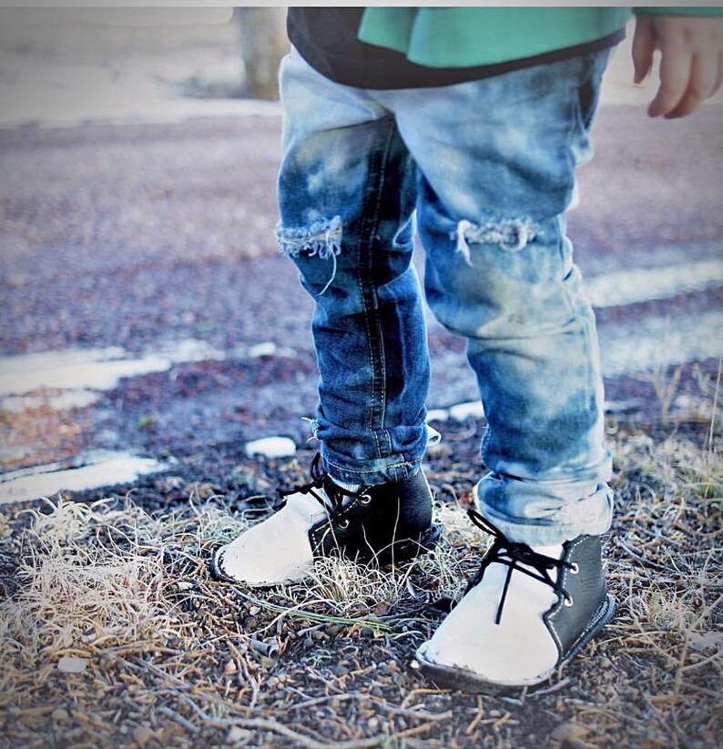 May include: A pair of black and white leather shoes with laces. The shoes have a white toe and a black heel. The shoes are worn by a person wearing ripped blue jeans.