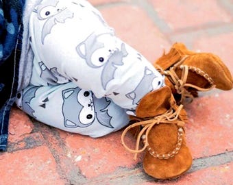 May include: A pair of brown suede baby moccasins with fringe and laces. The moccasins are worn by a baby wearing grey and white leggings with a cartoon animal print.