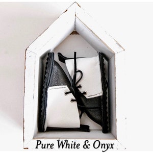 May include: A pair of black and white leather baby boots with laces. The boots are in a white wooden house-shaped box. The text "Pure White & Onyx" is below the box.