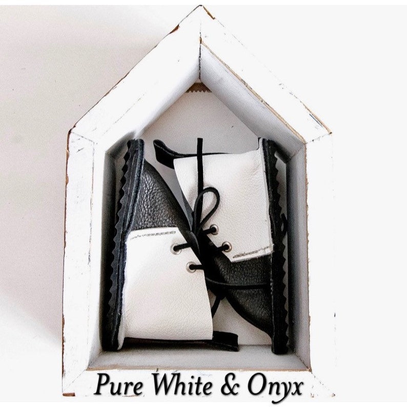 May include: A pair of black and white leather baby boots with laces. The boots are in a white wooden house-shaped box. The text "Pure White & Onyx" is below the box.