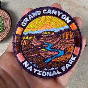 Puede incluir: Parche bordado con una colorida representación del Parque Nacional del Gran Cañón. El parche circular tiene un borde morado con las palabras "GRAND CANYON NATIONAL PARK" en blanco. La imagen muestra el cañón, un río y un amanecer.