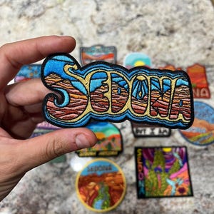 May include: Embroidered patch with the word "SEDONA" in a stylized font. The patch features a landscape design with blue, orange, and brown colours, and a blue border. Other patches with landscape designs are visible in the background.