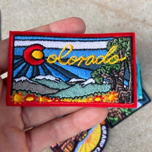 Puede incluir: Parche bordado con la palabra "Colorado" en escritura amarilla, un sol rojo y un paisaje montañoso. El parche tiene un borde rojo y está diseñado para aplicarse en ropa o bolsos.