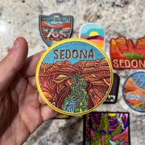 May include: A selection of embroidered patches with landscape designs. The main patch reads "SEDONA" in blue, above a detailed canyon scene with a river. Other patches depict desert landscapes and nature themes.