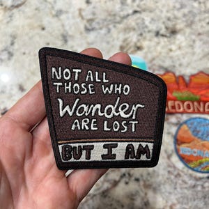 Può includere: Toppa ricamata marrone con la scritta "NOT ALL THOSE WHO Wander ARE LOST BUT I AM" in lettere bianche. La toppa ha un bordo nero e una forma unica.