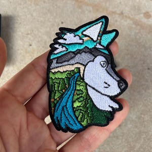 以下が含まれることがあります： 風景デザインのオオカミの頭部が刺繍されたパッチ。パッチには滝、山、ターコイズブルーの空が含まれています。オオカミの頭部はグレーと白で、黒い縁取りがあります。
