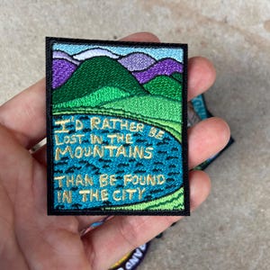 Puede incluir: Parche bordado con un paisaje de montaña con colinas verdes, montañas moradas y un lago azul. El parche tiene el texto "I'd rather be lost in the mountains than be found in the city" en letras doradas.