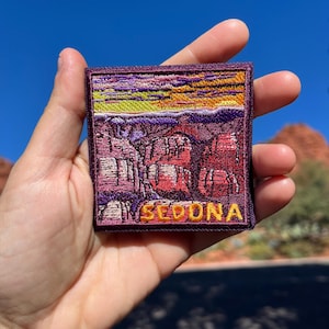 Puede incluir: Parche bordado con un paisaje de Sedona, Arizona, con formaciones rocosas rojas bajo un cielo de puesta de sol. El parche tiene un borde morado y la palabra "SEDONA" en letras doradas.