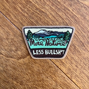 Può includere: Toppa ricamata con una scena naturalistica con montagne, alberi e un lago. La toppa ha il testo "More Nature, Less Bullsh*t" in verde e bianco. La toppa è bordata in marrone e beige.