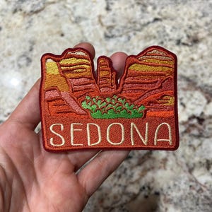 May include: Embroidered patch featuring a desert landscape with red rock formations and green shrubbery. The word "SEDONA" is embroidered in a serif font at the bottom of the patch. The patch has a red border.