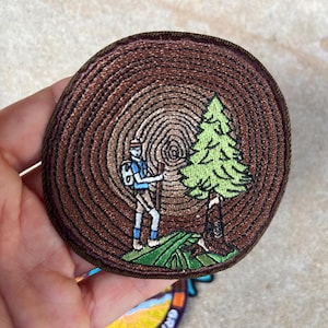 Könnte beinhalten: Runder, gestickter Aufnäher mit einem Wanderer mit Rucksack und Wanderstock neben einer grünen Kiefer. Der Hintergrund ist ein braunes Baumringmuster. Der Wanderer trägt blaue und weiße Kleidung.