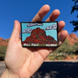 Puede incluir: Parche bordado con Bell Rock en Sedona, Arizona. El parche muestra una formación rocosa roja bajo un cielo azul con nubes blancas. El texto "BELL ROCK SEDONA" está bordado en la parte inferior.