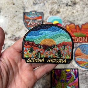 Può includere: Toppa ricamata raffigurante un paesaggio desertico con cactus, formazioni rocciose rosse e un tramonto sotto un cielo blu. La toppa è semicircolare con la scritta "SEDONA ARIZONA" in lettere dorate.