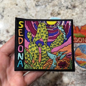 Puede incluir: Parche bordado con un diseño de paisaje desértico colorido, con un cactus, colibríes y un sol poniente. La palabra "SEDONA" está bordada verticalmente en el lado izquierdo. Borde negro y colores vibrantes.