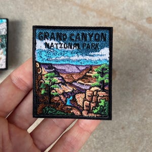 Può includere: Toppa ricamata raffigurante il Parco Nazionale del Grand Canyon. La toppa ha un bordo nero e il testo "GRAND CANYON NATIONAL PARK" in blu e bianco. L'immagine raffigura il canyon con sfumature di blu, viola, verde e marrone.