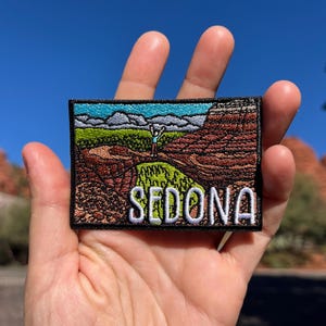 May include: Embroidered patch featuring a landscape scene with the word "SEDONA" in white block letters. The patch depicts a canyon with red rock formations, green vegetation, and a blue sky. The patch is rectangular with a black border.