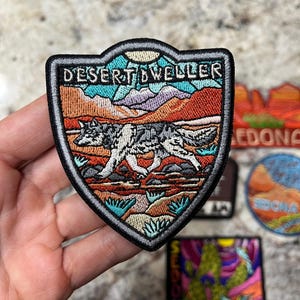 Può includere: Toppa ricamata a forma di scudo, raffigurante un lupo che attraversa un paesaggio desertico. La toppa ha un bordo nero e la scritta "DESERT DWELLER" in alto. Lo sfondo comprende montagne, un sole e vari colori.