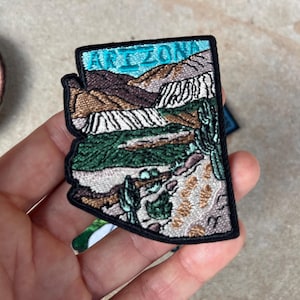 Puede incluir: Parche bordado con la forma de Arizona, que representa un paisaje con montañas, cactus y la palabra "ARIZONA" en turquesa. El parche tiene un borde negro y está sostenido en una mano.