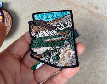 Puede incluir: Parche bordado con la forma de Arizona, que representa un paisaje con montañas, cactus y la palabra "ARIZONA" en turquesa. El parche tiene un borde negro y está sostenido en una mano.
