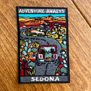 Può includere: Toppa ricamata con la scritta "ADVENTURE AWAITS" sopra un colorato paesaggio desertico con una strada e un veicolo. La parola "SEDONA" è ricamata sotto. La toppa ha un bordo nero.