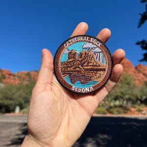 May include: A circular patch with the words "CATHEDRAL ROCK SEDONA" around a landscape image. The patch shows red rock formations, a blue sky, and a brown border. The patch is held in a hand against a blue sky.