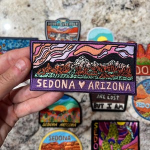 May include: Embroidered patches with desert landscape designs. One patch features the text "SEDONA ARIZONA" with a heart symbol. Other patches display desert scenes and the word "SEDONA". The patches have vibrant colors and detailed stitching.