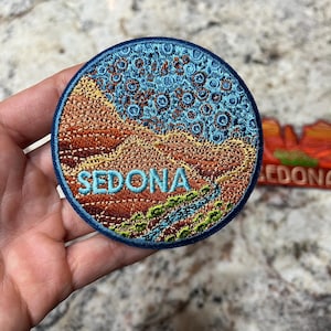 May include: Circular embroidered patch with a blue border, featuring a landscape design and the word "SEDONA". The patch depicts a desert scene with red rock formations, a blue sky, and a green river. The patch is round and suitable for clothing or bags.