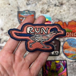 May include: Embroidered patch featuring a roadrunner bird with the word "RUN" above it. The patch has a pink background, brown border, and blue accents. Other patches are visible in the background.