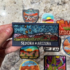 May include: A selection of embroidered patches with desert landscape designs and the text "Sedona Arizona." The patches display vivid colours and intricate stitching, with images of cacti, sunsets, and a wolf. The patches are rectangular, circular, and shield-shaped.