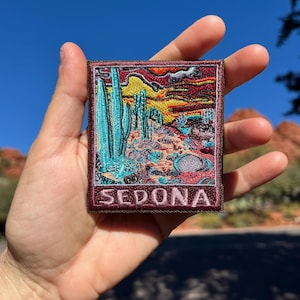 Puede incluir: Parche bordado con un paisaje desértico con cactus turquesas, una colorida puesta de sol y la palabra "SEDONA". El parche tiene un borde granate y es de forma cuadrada.