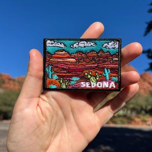 May include: An embroidered patch depicting a desert scene with red rock formations, cacti, and the word "SEDONA." The patch has a black border, a turquoise sky with white clouds, and is held in a hand against a blue sky. The patch is approximately 7.6 cm x 5 cm.