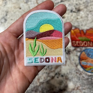 May include: Embroidered patch featuring a desert landscape with a yellow sun, mountains, and cacti. The patch has the word "SEDONA" in colorful letters at the bottom. The patch is rectangular with rounded corners and a white border.