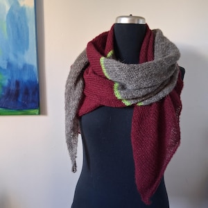 Könnte beinhalten: Gestrickter Schal in Burgunder- und Grautönen, drapiert auf einer schwarzen Schaufensterpuppe. Der Schal hat einen kontrastierenden neongrünen Streifen am Rand. Der Schal ist aus weichem, strukturiertem Garn gefertigt.