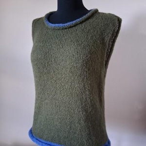 Könnte beinhalten: Olivgrüne Strickweste mit blauem Besatz am Halsausschnitt und Saum. Das ärmellose Oberteil wird auf einer schwarzen Schaufensterpuppe präsentiert. Die Strickstruktur ist sichtbar, und der blaue Besatz setzt einen Kontrast.