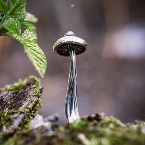 May include: A silver mushroom sculpture with a detailed, textured stem. The sculpture is set against a blurred background of green foliage.
