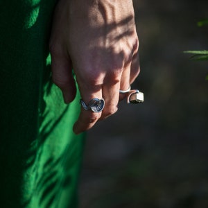May include: A person's hand wearing two silver rings. The ring on the index finger is a detailed, organic design. The ring on the middle finger is a simple band with a square-shaped stone.