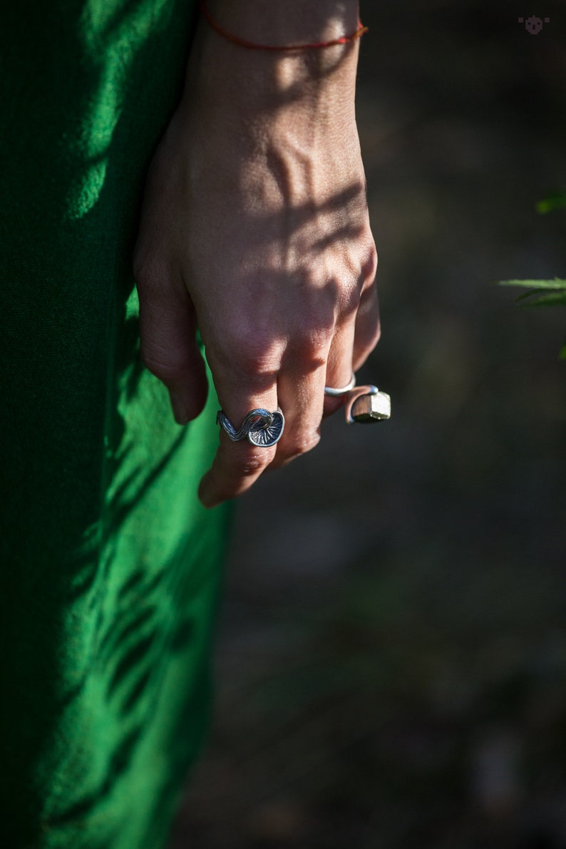 May include: A person's hand wearing two silver rings. The ring on the index finger is a detailed, organic design. The ring on the middle finger is a simple band with a square-shaped stone.