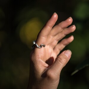 May include: A silver ring with a wavy design. The ring is worn on the middle finger of a person's hand.
