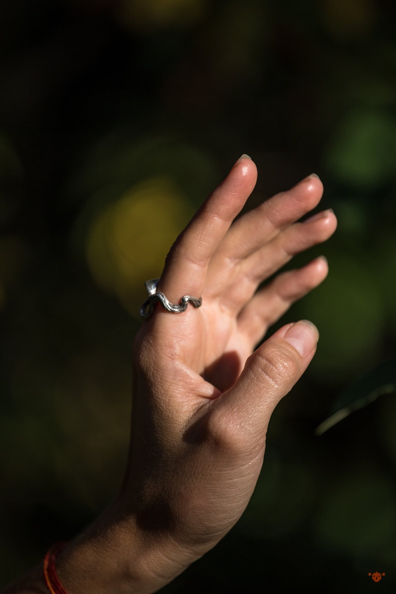 May include: A silver ring with a wavy design. The ring is worn on the middle finger of a person's hand.