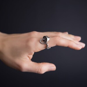 May include: A silver ring with a unique design featuring a curved, abstract shape. The ring is worn on a finger and is a statement piece of jewelry.