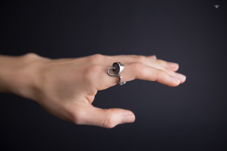 May include: A silver ring with a unique design featuring a curved, abstract shape. The ring is worn on a finger and is a statement piece of jewelry.