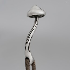 May include: A silver mushroom charm with a detailed stem and cap. The charm is attached to a brown cord.
