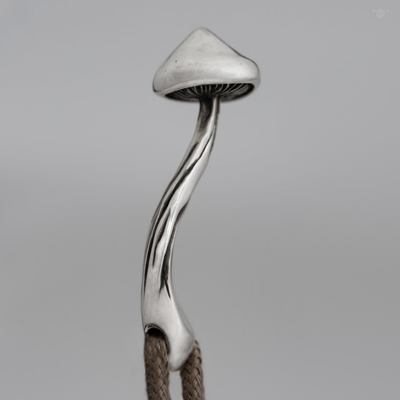 May include: A silver mushroom charm with a detailed stem and cap. The charm is attached to a brown cord.