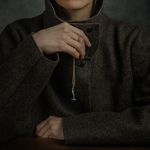 May include: A person wearing a brown wool jacket with a brown leather cord and a silver pendant. The person is holding the cord with their right hand.