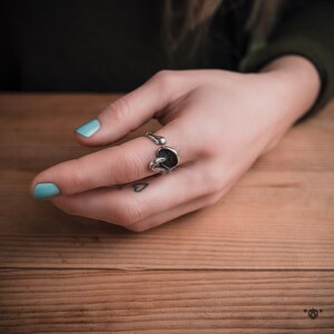 May include: A silver ring with a unique design featuring a circular element and a curved shape. The ring is worn on a finger with light blue nail polish.