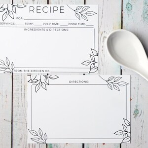 May include: Two white recipe cards with black leaf corner designs and the word "RECIPE" at the top. The cards have spaces for ingredients, directions, and other details. A white spoon rests on a light wood surface.