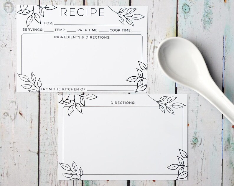 May include: Two white recipe cards with black leaf corner designs and the word "RECIPE" at the top. The cards have spaces for ingredients, directions, and other details. A white spoon rests on a light wood surface.